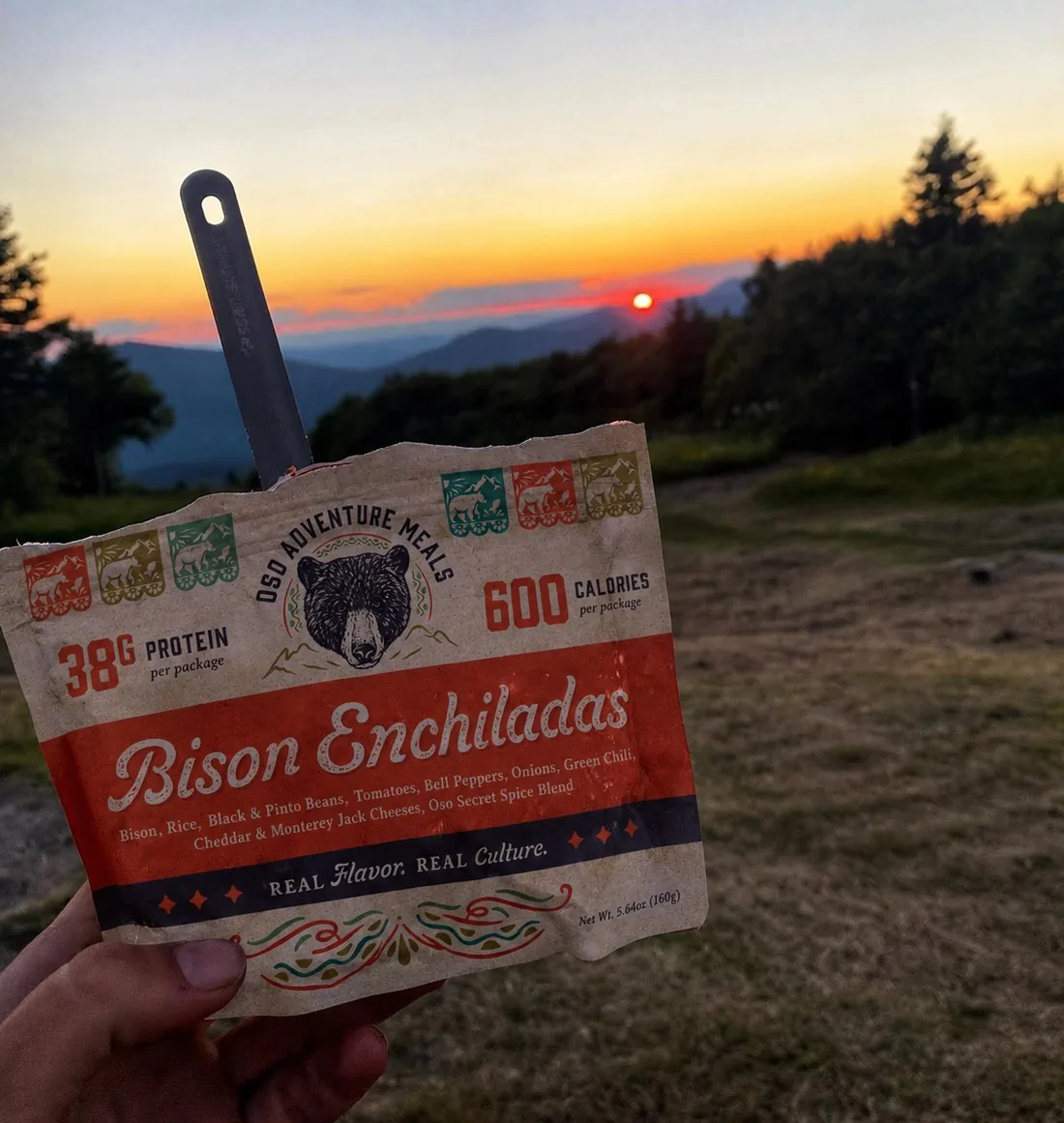 A Bison Enchiladas pouch with a spoon in it, held up at sunset over the mountains