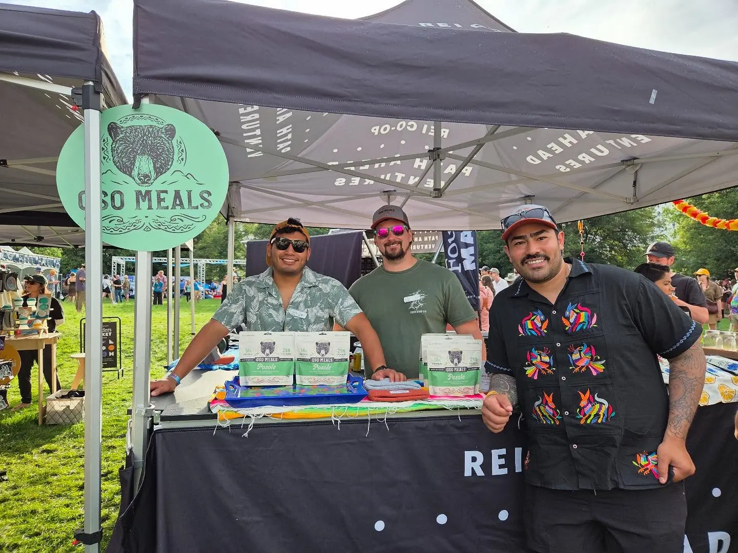 Three of the Oso crew smiling behind their booth under the Oso Meals sign at an outdoor festival