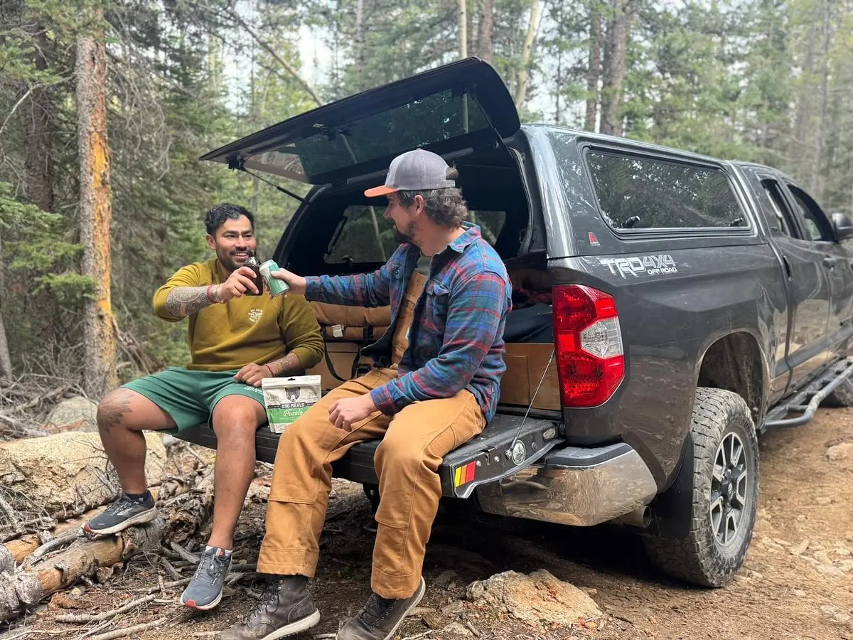 Two friends raising a drink on a truck tailgate in the woods with an Oso Posole pouch